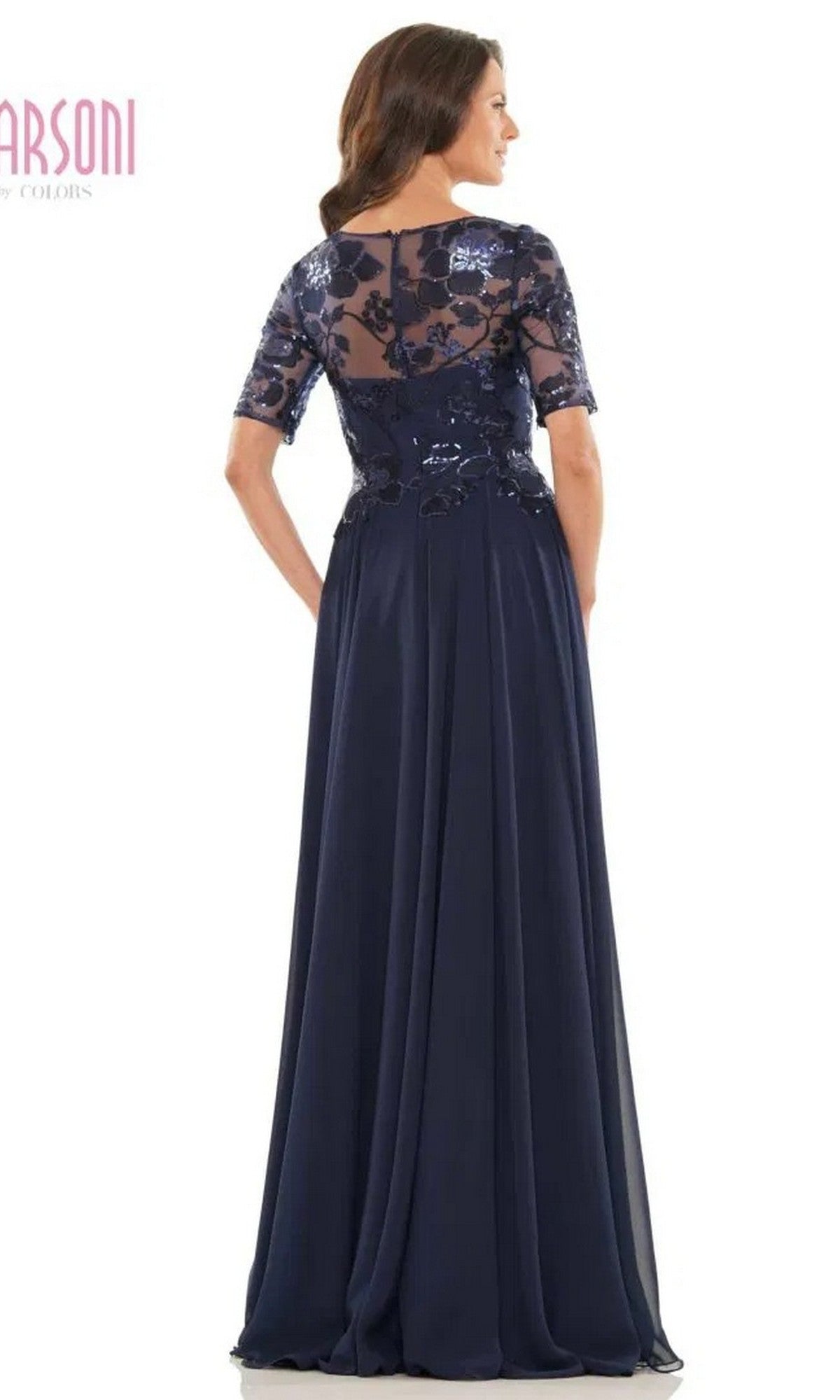 A woman stands with her back to the camera in the Marsoni M286 long formal dress, a navy blue gown featuring a sequin lace bodice, sheer embroidered short sleeves, and a sheer back panel.
